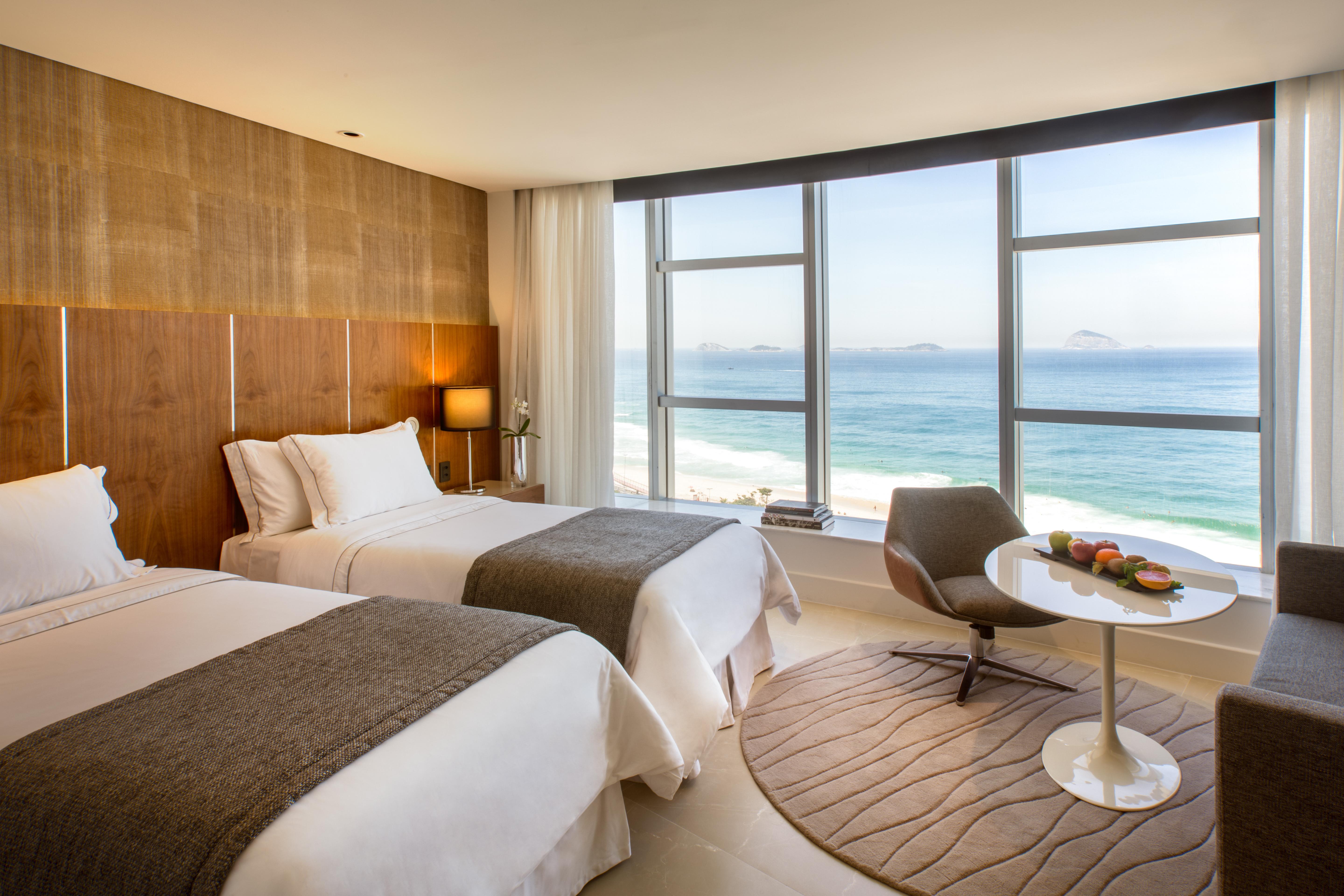 Hotel Nacional Rio De Janeiro - Oficial Kültér fotó The photo shows a modern hotel room with two beds adorned in white linens and gray throws. Large windows allow natural light to flood the space, offering a stunning view of the ocean. In the corner, there's a comfortable chair and a small round table